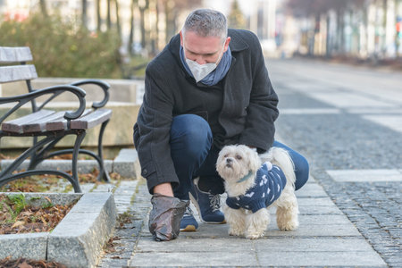 Mature man wearing a protective mask collects dog poop into a bag while walking down a city street