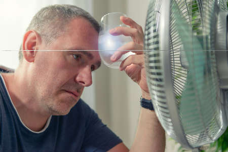 Man suffers from heat while sitting in the oom and tries to cool off by the fanの写真素材