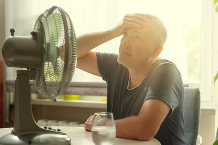 Man suffers from heat while sitting in the oom and tries to cool off by the fanの写真素材