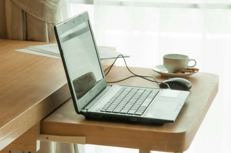 Close up view of a work desk interior with a laptop computer, a cup of coffee and white curtainsの写真素材