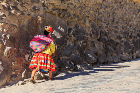 Peruvian woman cholita dressed in traditional colorful cloth, carrying the sack and walking up the street with stony walls, Inkan Sacred Valley, Ollantaytambo, Peruの写真素材
