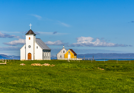Flateyjarkirkja white lutheran church and couple of living huts with meadow in foreground and sea  fjord with blue sky in the background, Flatey, Icelandの写真素材