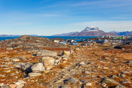 Autumn greenlandic  tundra with orange grass, stones, Inuit settlement and Sermitsiaq mountain in...