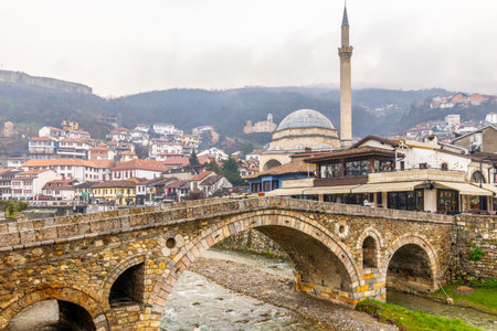 Stone arch bridge crossing a shallow Bistrica river with Ottoman Sinan Pasha mosque and tall minaret rising above dense hillside housing and traditional architecture on overcast day, Prizren, Kosovoの写真素材