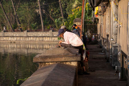 Kolhapur, India- December 5th 2020; Stock photo of 50 to 60 year old Indian grandpa or man wearing casual cloths and blue cap enjoying fishing at Rankala lake at early morning under bright sunlight.のeditorial素材