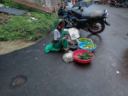 Kolhapur,India- July 14th 2021; Stock photo of 50 to 60 year old Indian grandma wearing green color traditional saree, sitting on the street for selling vegetable and counting money at Kolhapur city.のeditorial素材