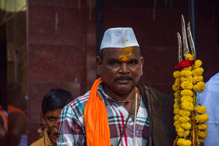 Yadgir,India- December 22nd 2019; Stock photo of Indian priest, wearing casual cloths, saffron color scarf,white cap and holding trishul decorated with marigold flower garland in Indian village templeのeditorial素材