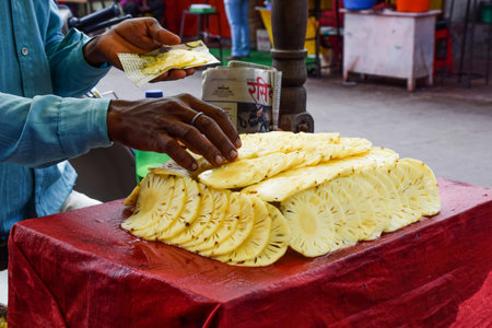 Tuljapur,India- December 19th 2019; Stock photo of fresh juicy sliced pineapple kept on big ice cube coverd with red cloth in wooden cart.Close up of fruit seller picking sliced pineapple for customerのeditorial素材