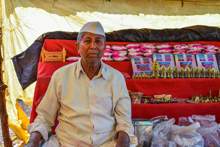 Kolhapur, India- January 31st 2019; Stock photo of 50 to 60 age old Indian grandpa wearing traditional white shirt , pant and cap sitting at his shop selling worshiping items. small shop on backgroundのeditorial素材
