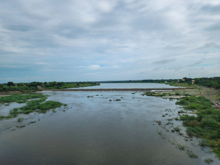 A serene countryside river flows under a bridge, reflecting a blue sky dotted with white clouds....
