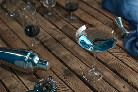 Cocktail in a martini glass blue on a wooden table next to a shaker and a jigger in the background of glassesの写真素材