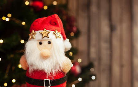 Christmas Santa toy in a red suit with golden stars on a hat and black belt on a Christmas tree background with a garland on a wooden backgroundの写真素材
