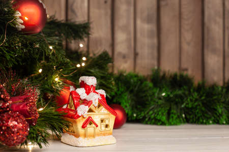 Christmas toy house on a light table next to a Christmas tree dressed up with red balls and a garland on a wooden backgroundの写真素材