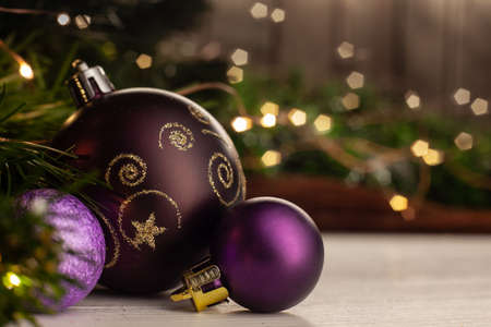 Christmas toy purple ball with a gold pattern next to small purple balls on a light table next to a Christmas tree on a background of a garland on a wooden wallの写真素材