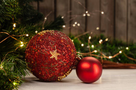 Christmas toy red ball decorated with beads and an asterisk next to a small red ball on a light table top next to a Christmas tree decorated with a garland against a wooden wallの写真素材