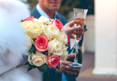 Wedding glasses with champagne in the hands of the newlyweds next to a wedding bouquet of white and scarlet rosesの写真素材