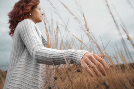 girl with red hair in a gray sweater walks across the field touching her ears of ears against the gray skyの写真素材