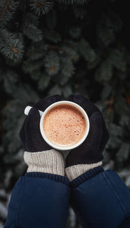 White cup of coffee in the hands dressed in gloves on the background of the Christmas tree on the streetの写真素材
