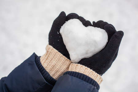 Hands in black gloves keep the snow folded in the shape of a heart against the background of snow outside.の写真素材