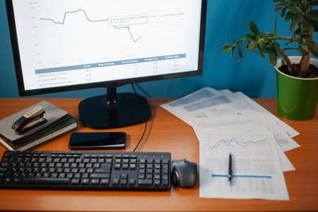 Desktop on which the printed chart is next to the keyboard and the daily on the background of the monitor with the image of graphs and a flower in a green potの写真素材