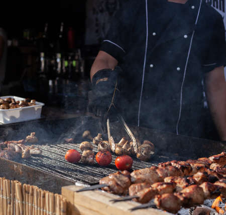 brazier in a black tunic grills meat and vegetables by turning them over with iron tongsの写真素材