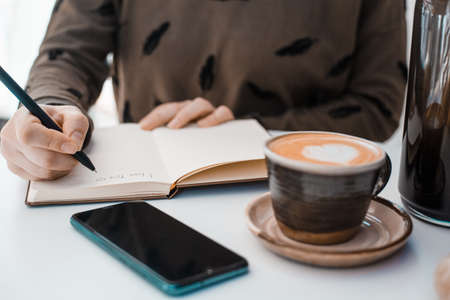 Girl writes I love you in a notebook on a table next to a phone and a cup of coffee decorated with a...