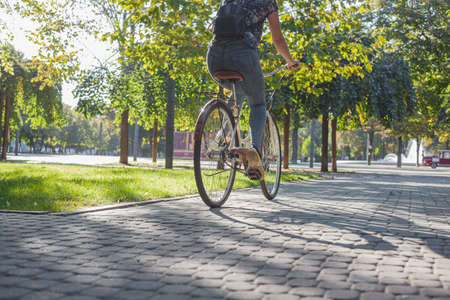A girl in jeans and sneakers rides a beige bike in the park among green trees and lawn