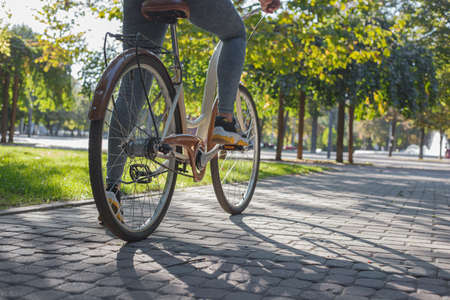 A girl in jeans and sneakers sits on a beige bicycle in the park among green trees and lawnの写真素材
