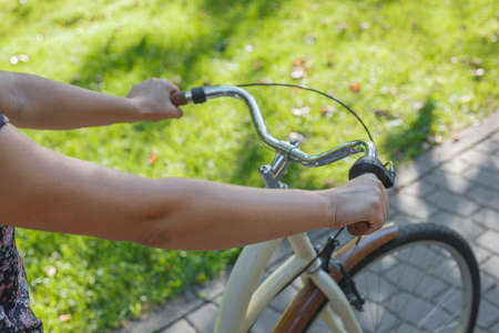 The girl holds the wheel of a beige bike in the park among green trees and lawnの写真素材