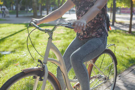 A girl in jeans and sneakers rides a beige bike in the park among green trees and lawn