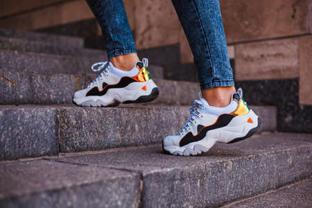 White sneakers with black inserts on female legs dressed in jeans on a marble staircase in the...
