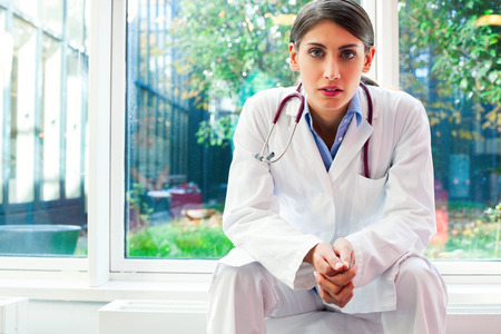 Portrait of serious female doctor with hands clasped. Young medical professional is wearing lab coat. She is sitting in hospital.の写真素材