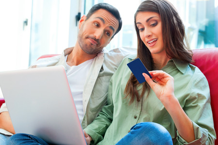 A photo of young man looking at woman with laptop and credit card. Male and female partners are in casuals. They are sitting on sofa at home.の写真素材