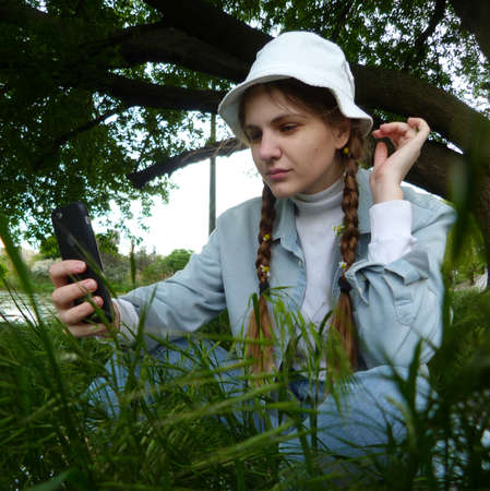 Beautiful girl photographing herself, sitting on green grass, with a tree on the background