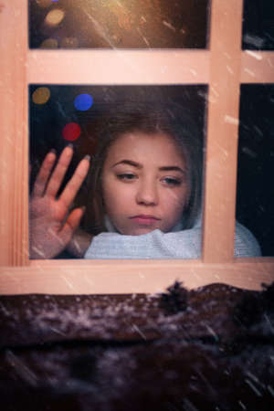 lonely and sad girl looks out the window in winter evening