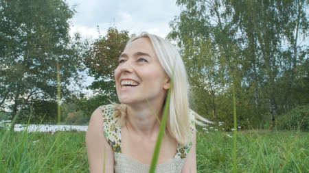 A young smiling beautiful woman in a light dress lies on the grass and looks at the sky against the background of trees.の写真素材