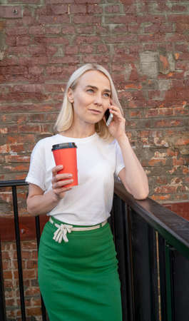 A beautiful girl talks on her mobile phone during lunchtime and drinks coffee from a red cup. A young girl drinks coffee from a green paper cup and looks away.の写真素材