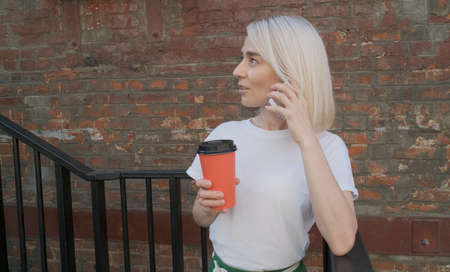A beautiful girl talks on her mobile phone during lunchtime and drinks coffee from a red cup. A young girl drinks coffee from a green paper cup and looks away.の写真素材