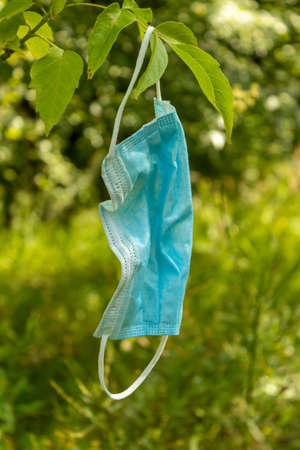 Waste during COVID-19. A used face mask hangs from a tree branch. Used medical face masks and gloves on the ground outdoor. Environmental pollution concept. Medical waste disposal. Copy spaceの写真素材