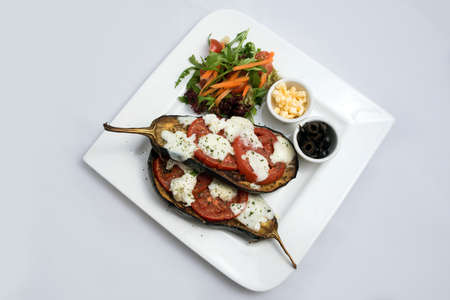 A low contrast warm Hero shot of a main course platter with grilled aubergine with buffalo cheese, tomato, olives & vegetables on the side, on a minimal white background with a 45 degree angle from diagonal perspective.の写真素材