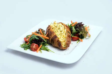 A low contrast image of a breakfast platter with egg-filled croissant with vegetables- carrot, tomato, lettuce on a minimal white background with a 30 degree angle from front perspectiveの写真素材