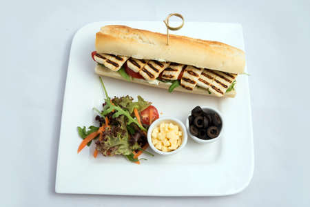A high contrast vertical Hero shot of a Grilled Halloumi cheese baguette with olives & salad on the side, on a minimal white background with a 90 degree angle from zoomed perspectiveの写真素材