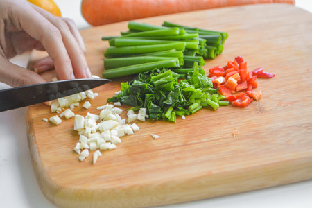 Female cook chopping fresh vegetables on wooden board: red and yellow bell pepper, green onions, chili, garlic, carrot and spinachの写真素材
