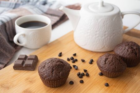 Chocolate muffins, chocolate chips, chocolate bar, cup of coffee, tea pot on a wooden surface with warm scarf, christmas lights bokeh and holly in the backgroundの写真素材