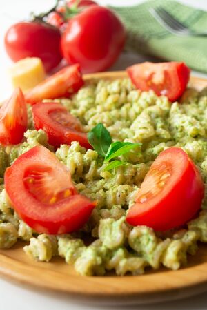 Basil pesto fusili pasta with Parmesan cheese and sliced tomatoes in a wooden plate and a green napkin with a fork in the backgroundの写真素材