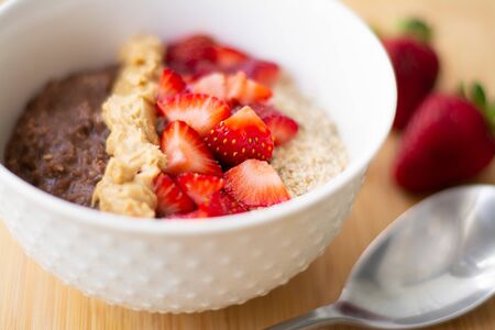 Chocolate cocoa oatmeal with almond meal, chopped strawberries and creamy peanut butter on a wooden surface. Strawberry, almonds, napkin and spoon in the backgroundの写真素材