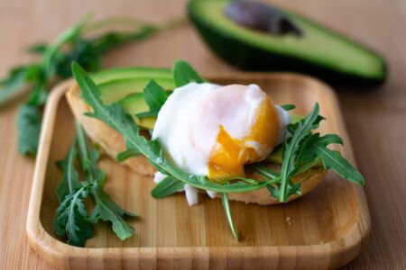 Stylish healthy breakfast of an avocado, arugula and poached egg toast on a wooden surface with half an avocado and arugula in the backgroundの写真素材