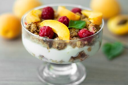 Healthy parfait with greek yoghurt, homemade granola, slices of apricot and raspberries in a glass goblet. Grey wooden background with apricots and basil leavesの写真素材
