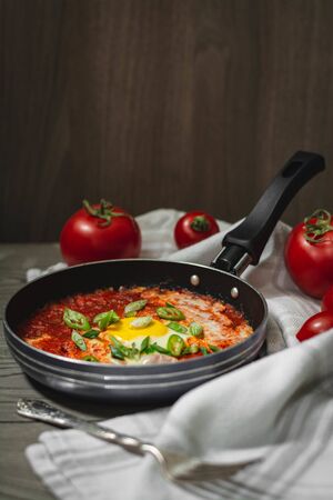 Shakshuka traditional middle eastern dish with tomatoes and eggs in a small pan garnished with scallions and chilli. Tomatoes in the background, dark and moody conceptの写真素材