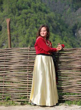 Photo of young brunette woman in a traditional georgian costume stands near a wicker fence on a background of mounains covered in a freen forestの写真素材
